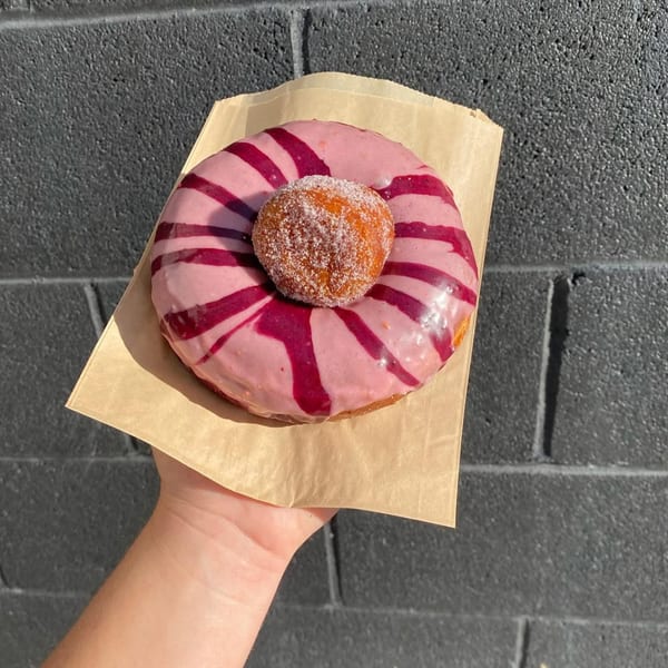 Hand holding a pink frosted donut with purple stripes and a sugar-dusted donut hole on top.