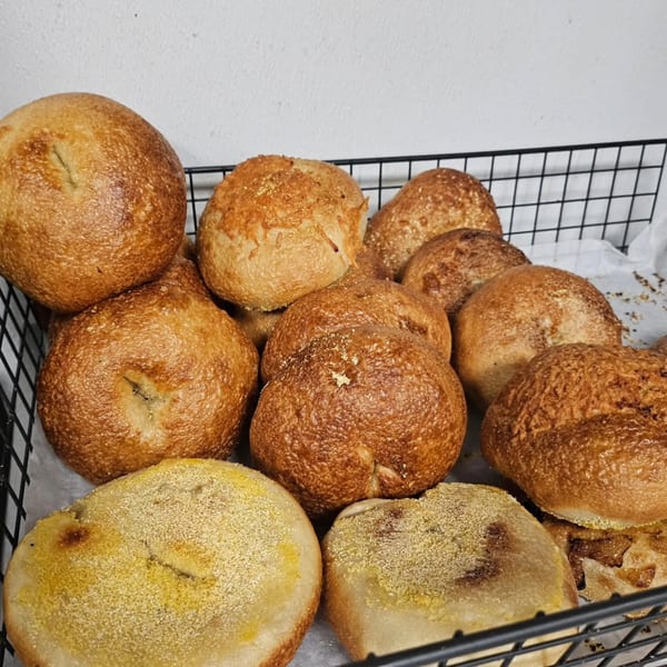 A black wire basket filled with various freshly baked bagels and English muffins from The Dough Miner.